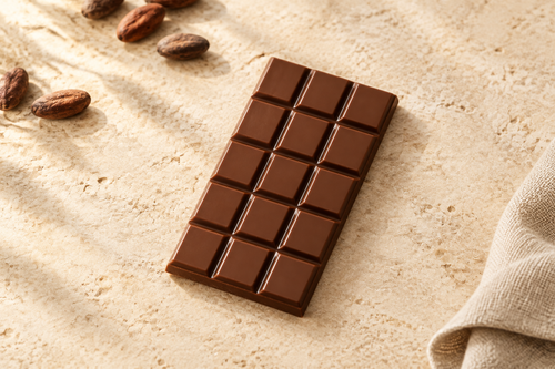 A rectangular chocolate bar with a 4-by-3 grid of dark brown squares, placed diagonally on a textured beige stone surface with cocoa beans and a folded linen cloth nearby.