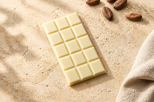 A white chocolate bar with a 3x5 grid of squares, placed diagonally on a textured beige stone surface with cocoa beans and a folded beige cloth nearby.